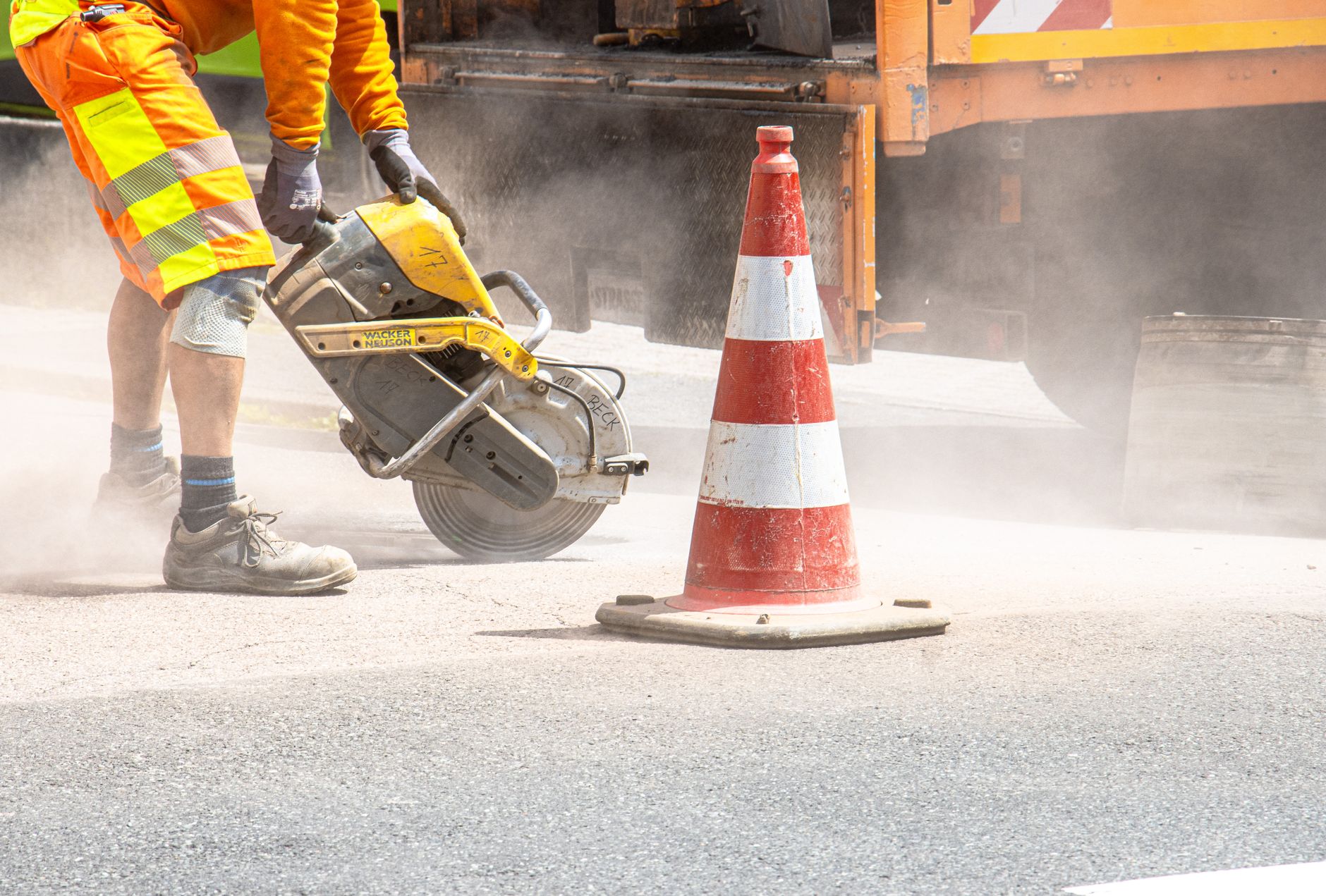 Ein Bauarbeiter schneidet mit einer großen Säge Asphalt neben einem Verkehrskegel auf einer Straße.