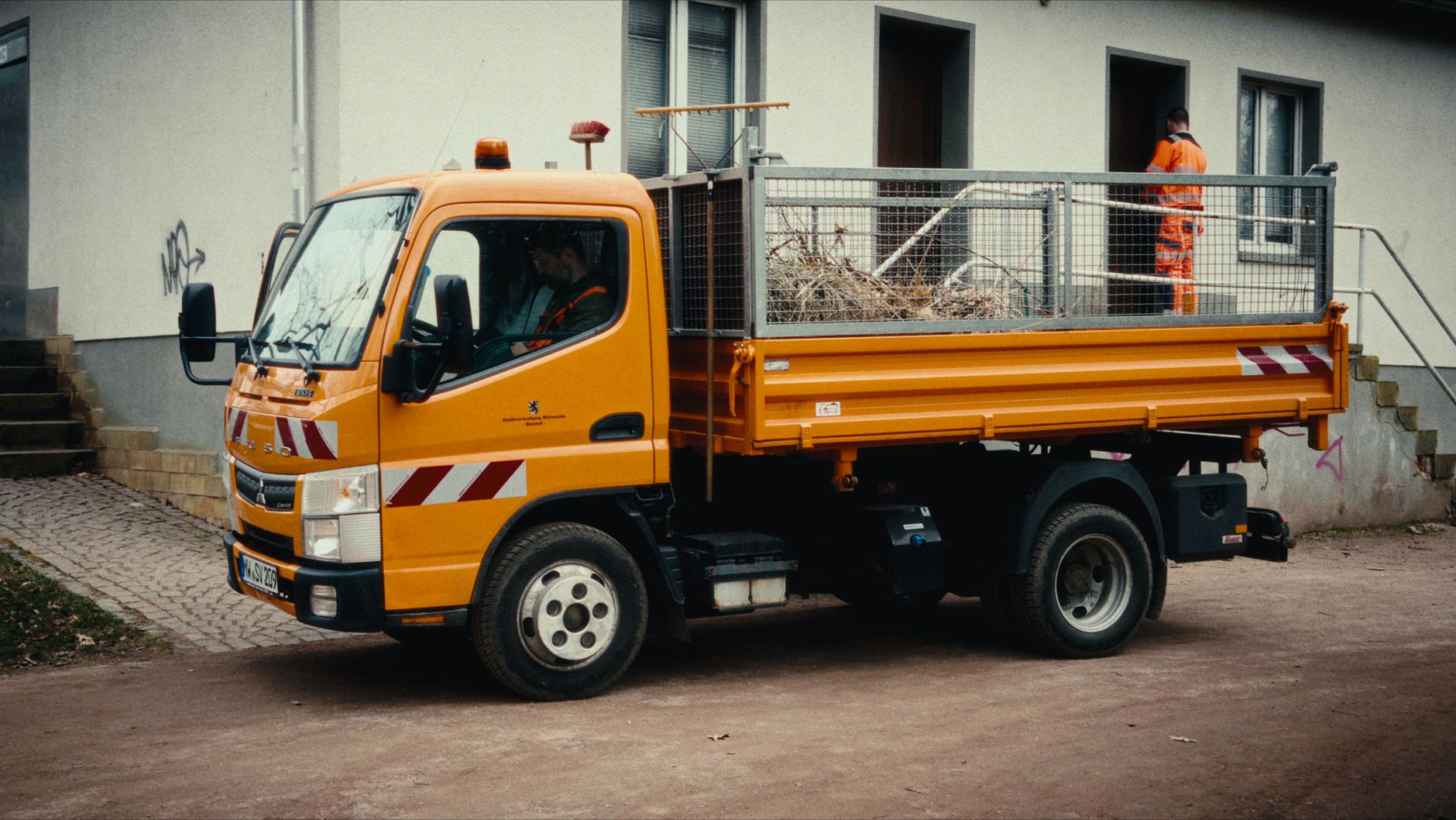 Orangefarbener Nutzlastwagen, der in der Nähe eines Gebäudes geparkt ist, mit Arbeitern in Warnkleidung.
