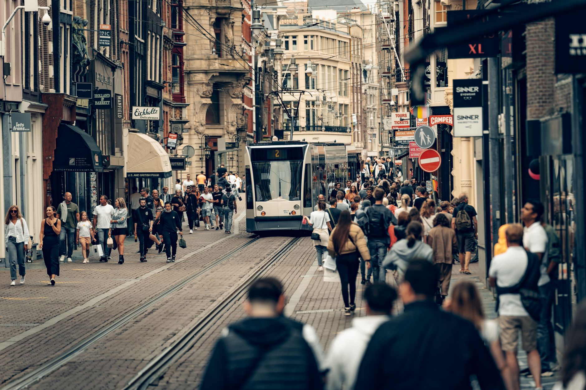 Geschäftliche Straßenszene in Amsterdam mit einer Straßenbahn, Geschäften und vielfältigen Fußgängern an einem lebhaften Tag.