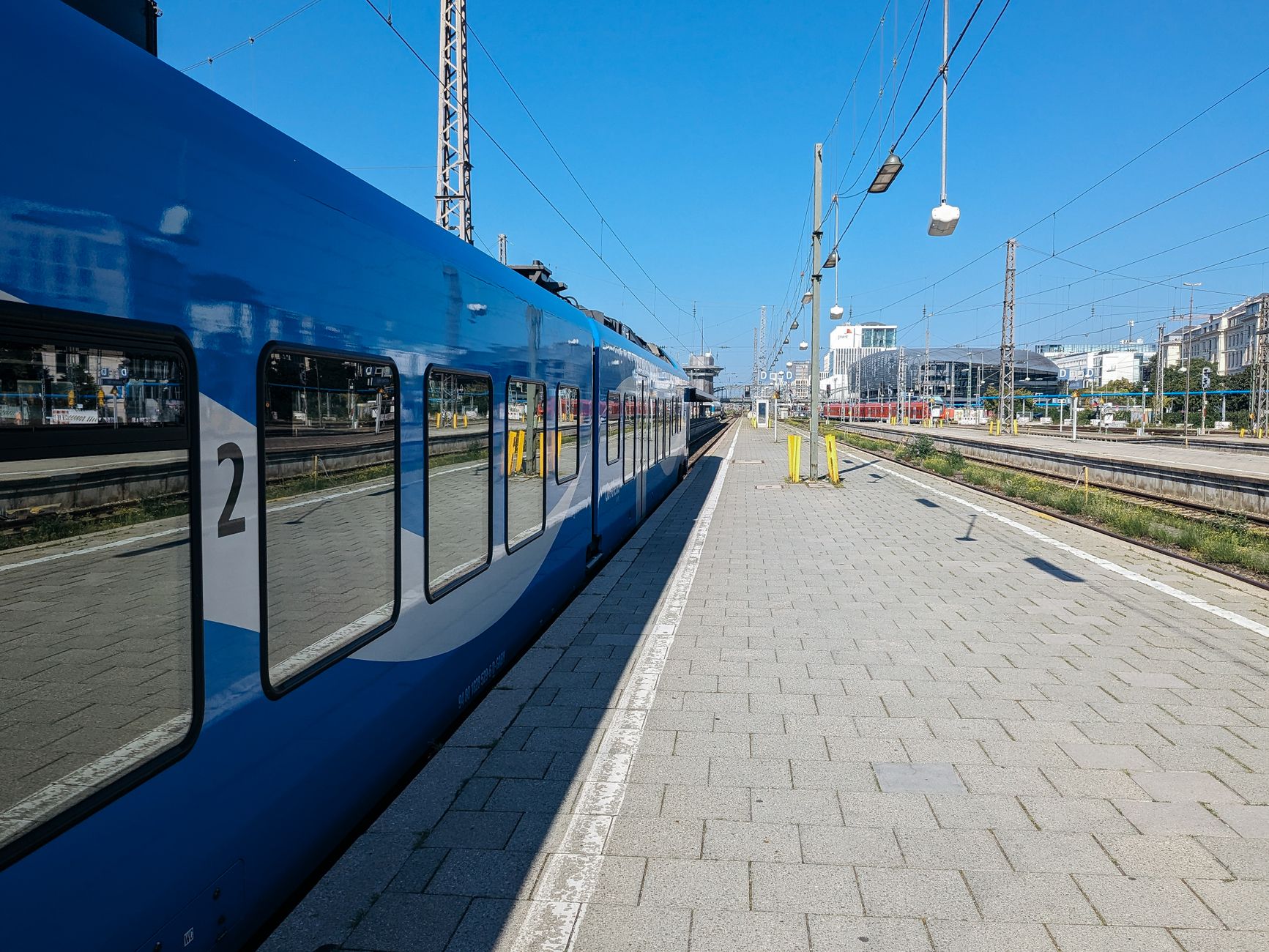 Ein blauer Zug auf einem Freiluftbahnsteig unter einem klaren Himmel in München.