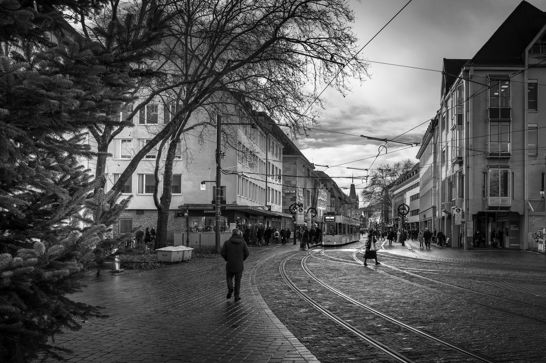 Schwarzweißaufnahme einer belebten Straße in Freiburg mit Straßenbahnschienen und Fußgängern im Herbst.