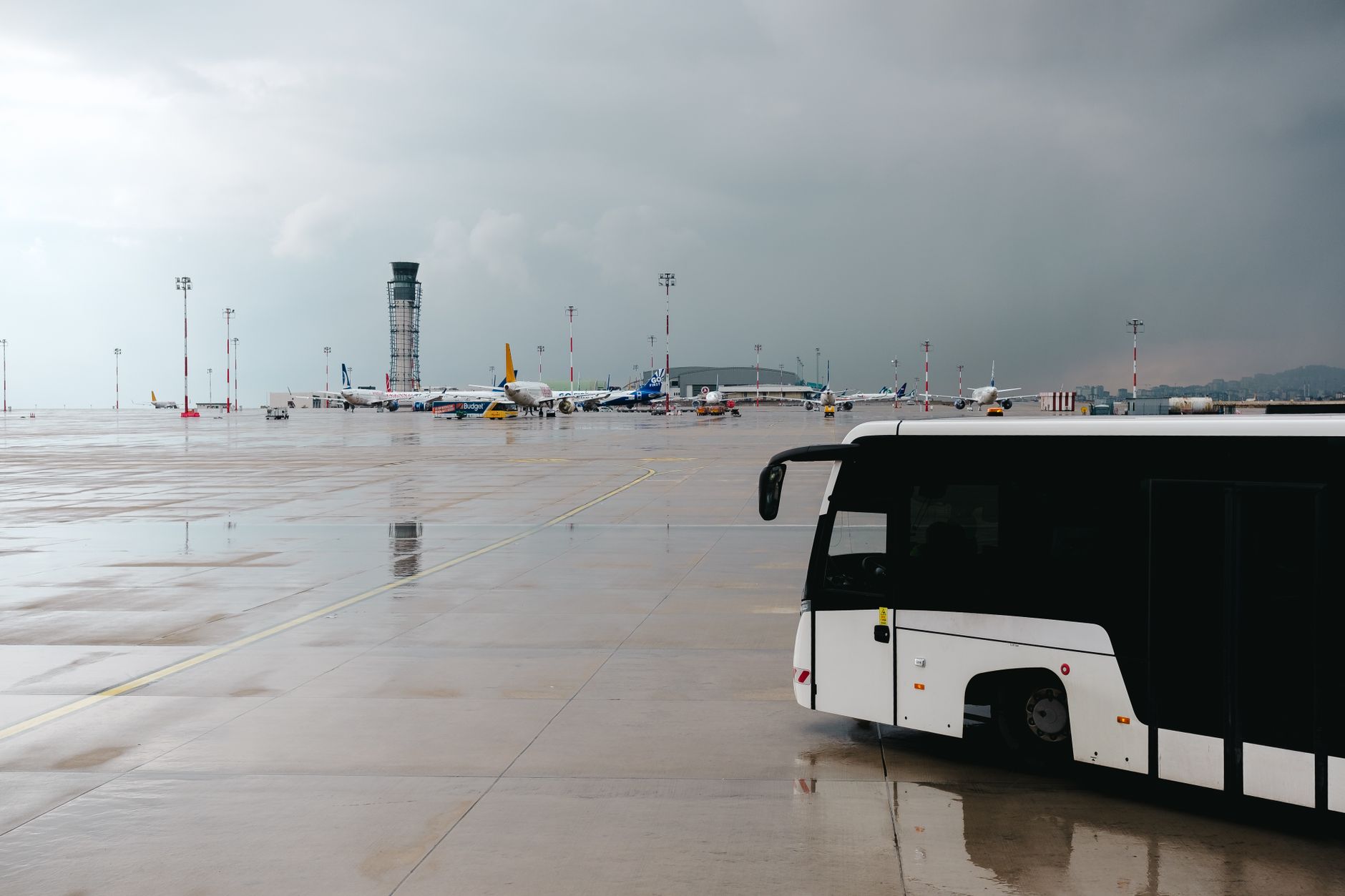 Verregnetes Flughafenrollfeld mit einem Passagierbus und Flugzeugen im Hintergrund.